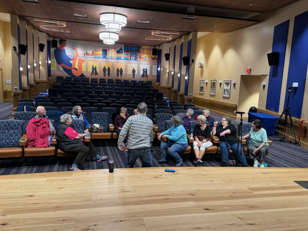 A man in a plaid shirt teaches improv to senior citizens in a small theater.